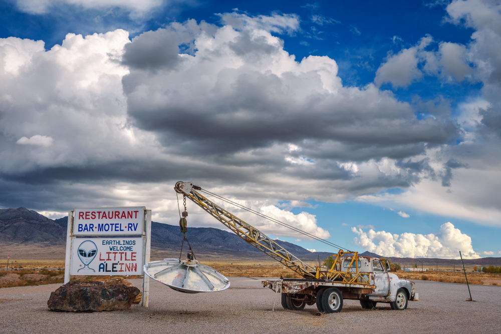 Sign for the Little A'le'inn and a tow truck holding a hanging UFO with big clouds overhead on the Extraterrestrial Highway.