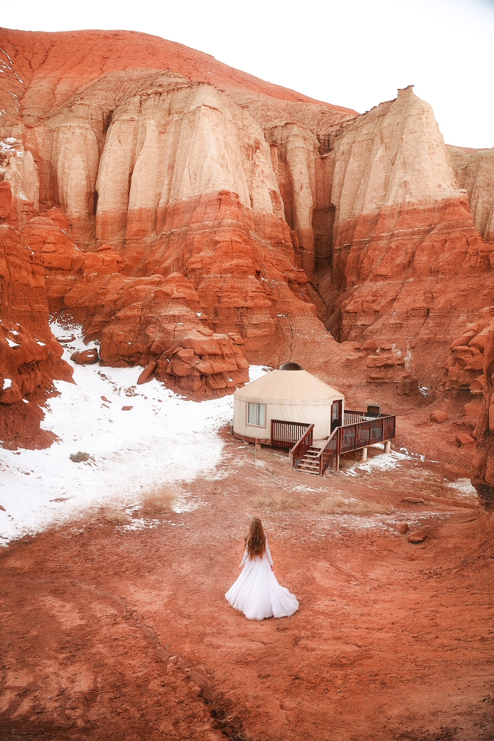 Woman with long hair and long white dress standing near a yurt and snow under striped rock cliff in Goblin Valley State Park.