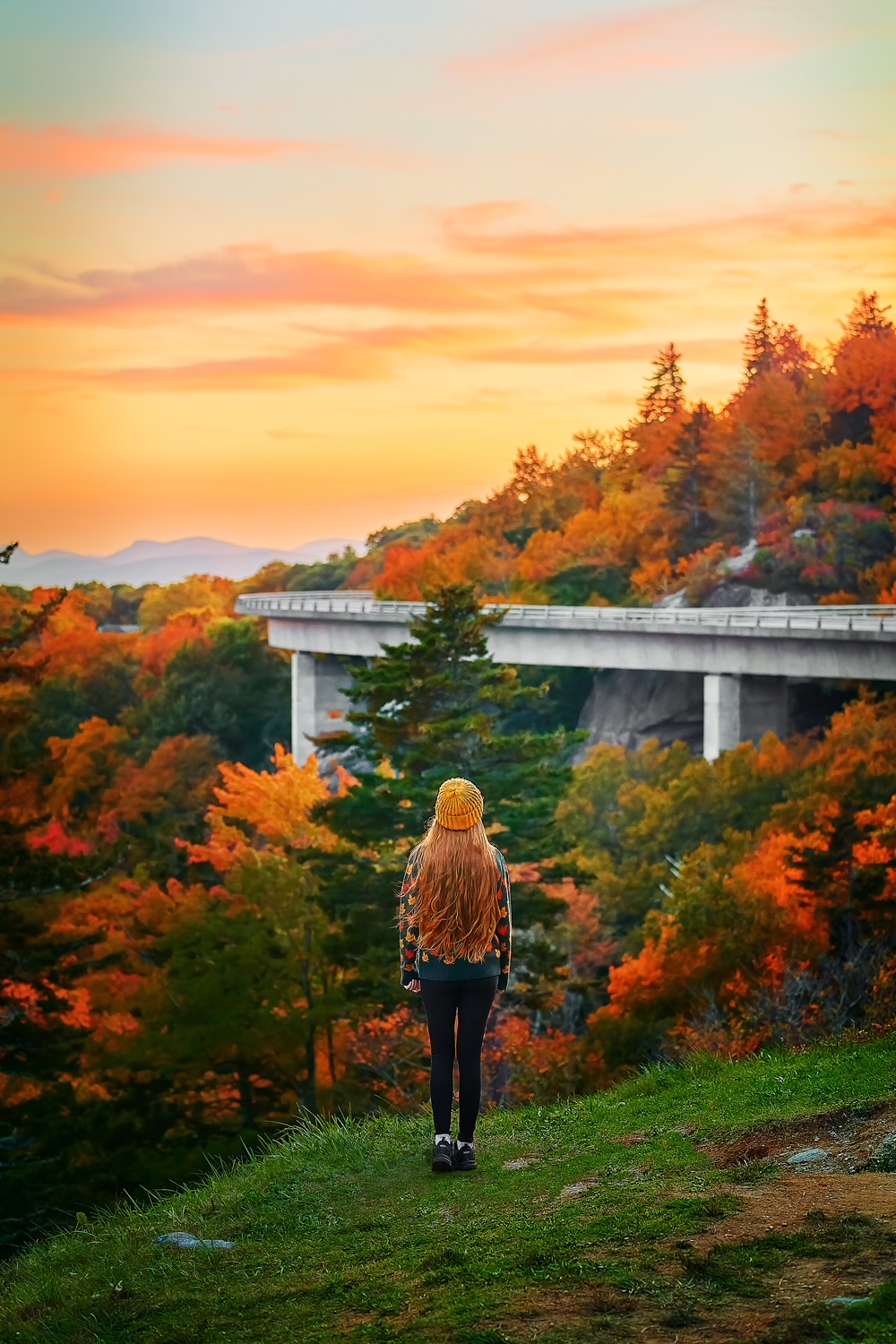 Woman with long hair and a yellow hat standing overlooking the Linn Cove Viaduct at sunset during the fall.