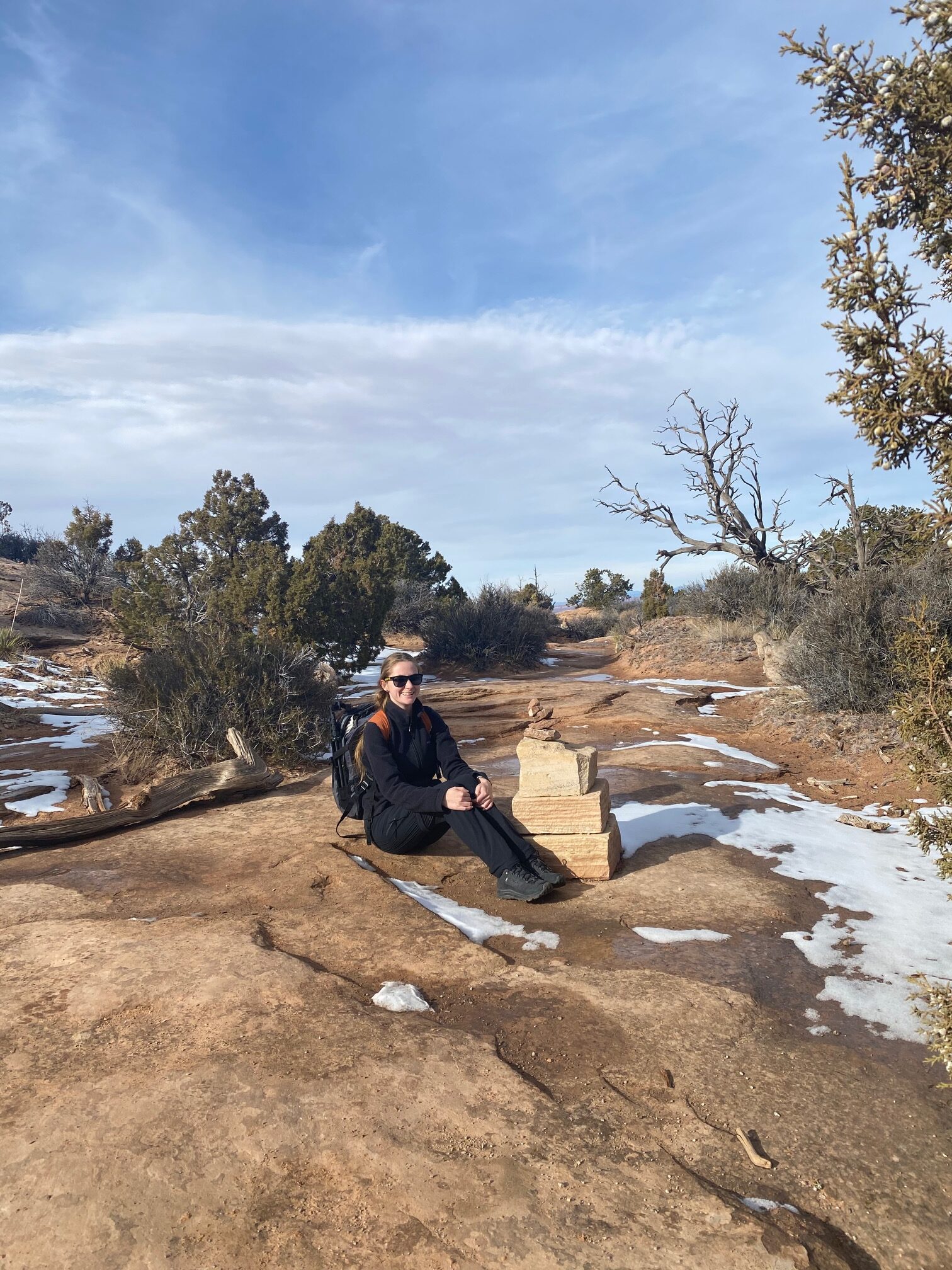 Woman in black and a backpack sitting on the ground next to a rock cairn among scrubby trees and a bit of snow.