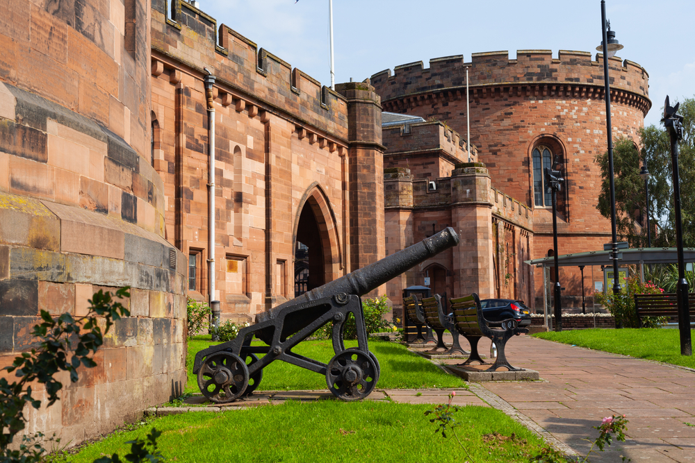 Castle with a courtyard with a gun in front of it. There are benches out the front. 