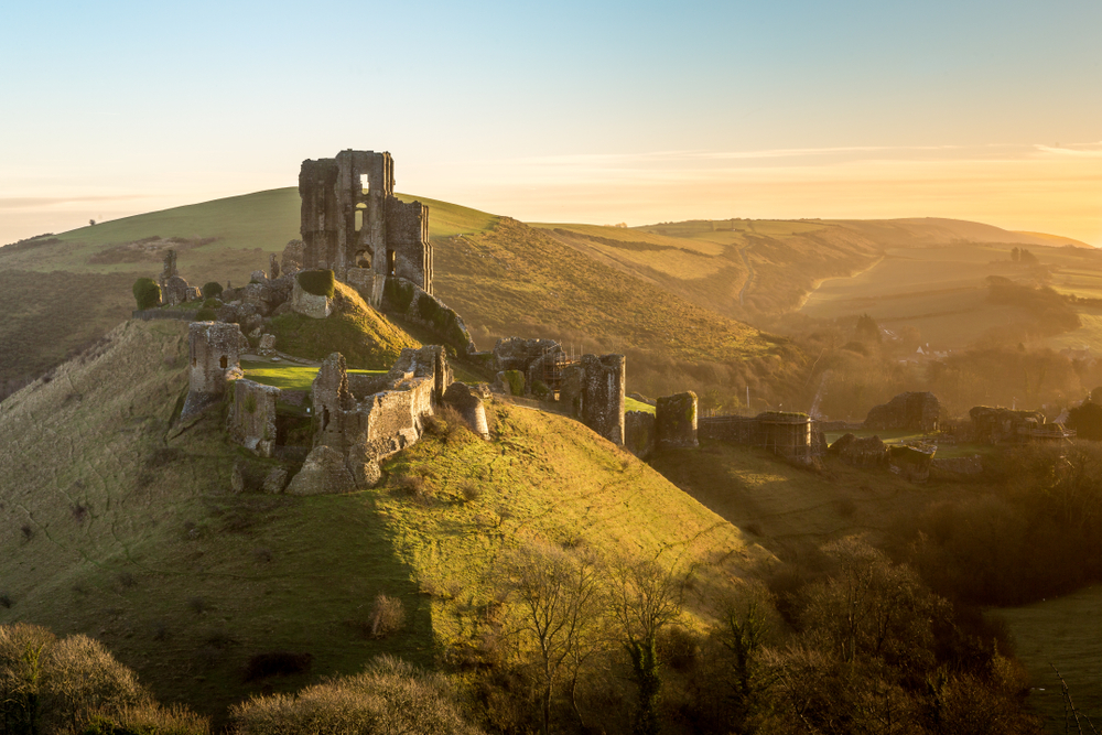 The ruins of Corfe Castle taken at sunset on top of a hill. The picture is taken from a distance and show the hill and all the castle. 