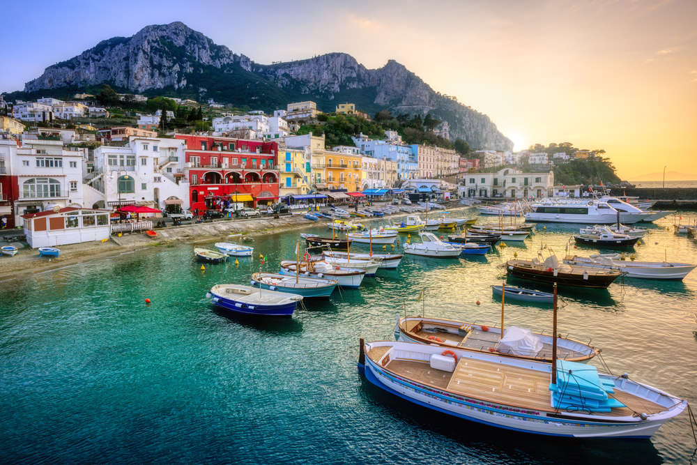 boats docked in the water off of the coast of capri, colorful houses are on the waters edge, mountains in the background in capri, one of the best places to visit in Europe in June 