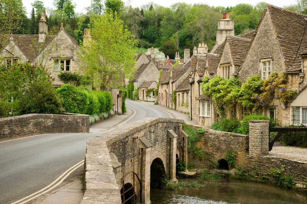 a road through a town of stone houses, there is a bridge over watering the foreground, trees in the background 