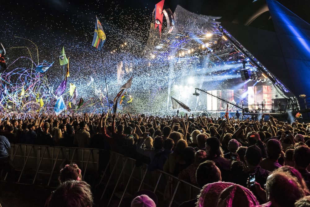 a large crowd of people in front of a stage as confetti falls from above and flags fly on tall poles, photo taken at night 