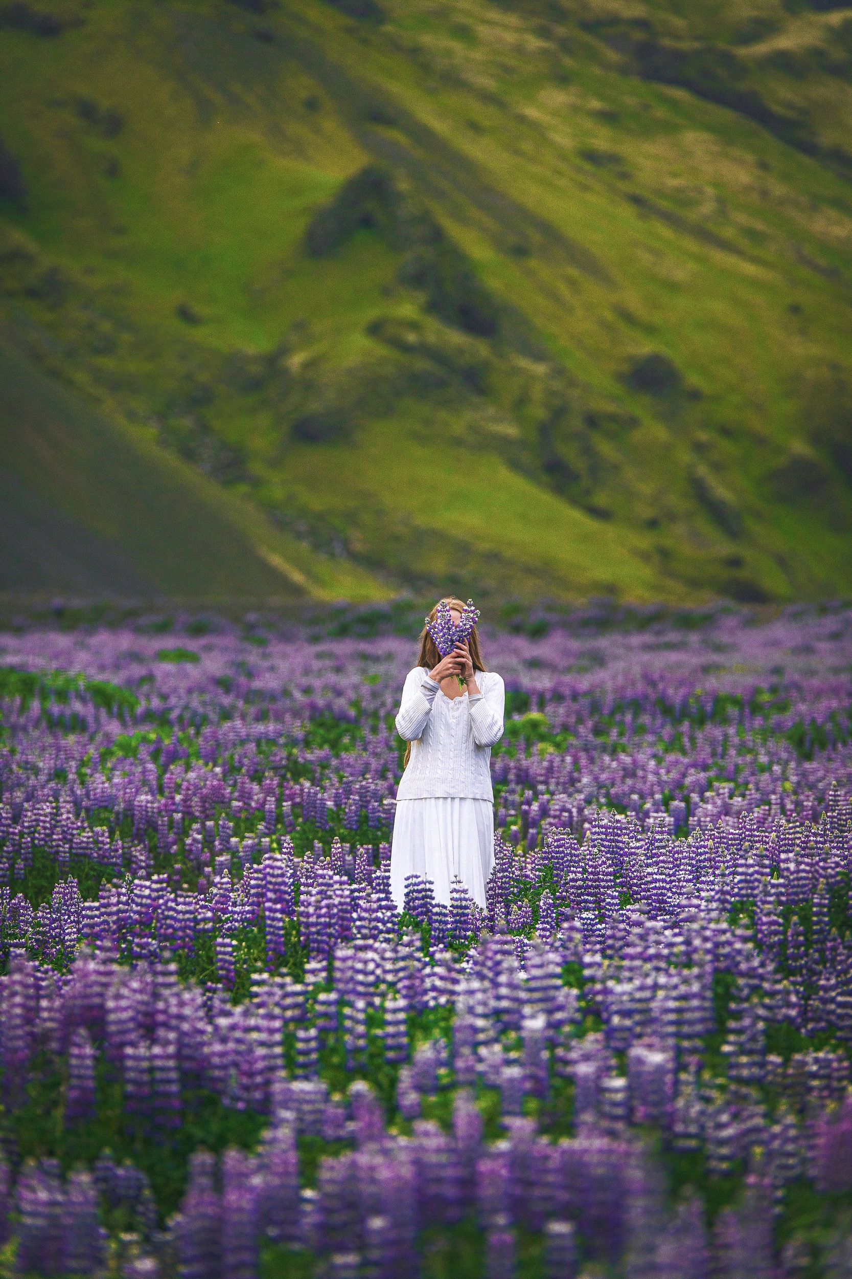 a woman in a sweater and a skirt is standing in a field of wildflowers in front of a mountain, she is holding some flowers in front of her face 