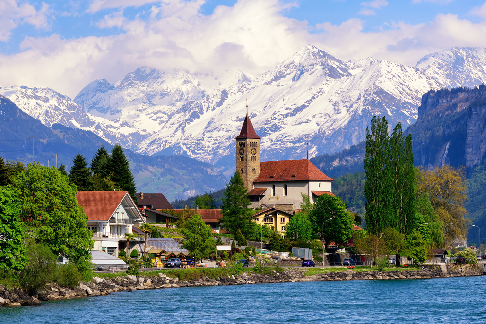 one of the best places to visit in Europe in June, the jungfrau region, snow covered mountains in the background and a river in the foreground with a town on the waters edge 