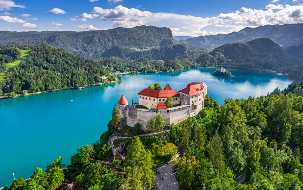 a castle next to a lake, the lake is bright blue and it is surrounded by trees and mountains 