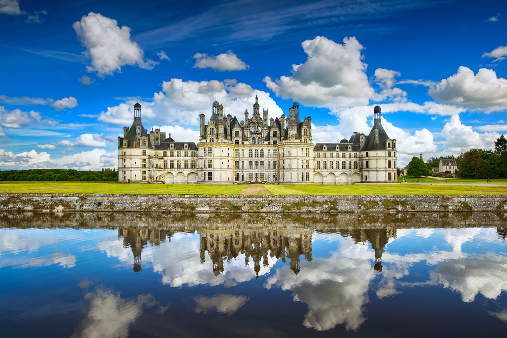 a large castle in front of a manicured lawn in front of a body of water, clouds in the sky, one of the best places to visit in Europe in June 