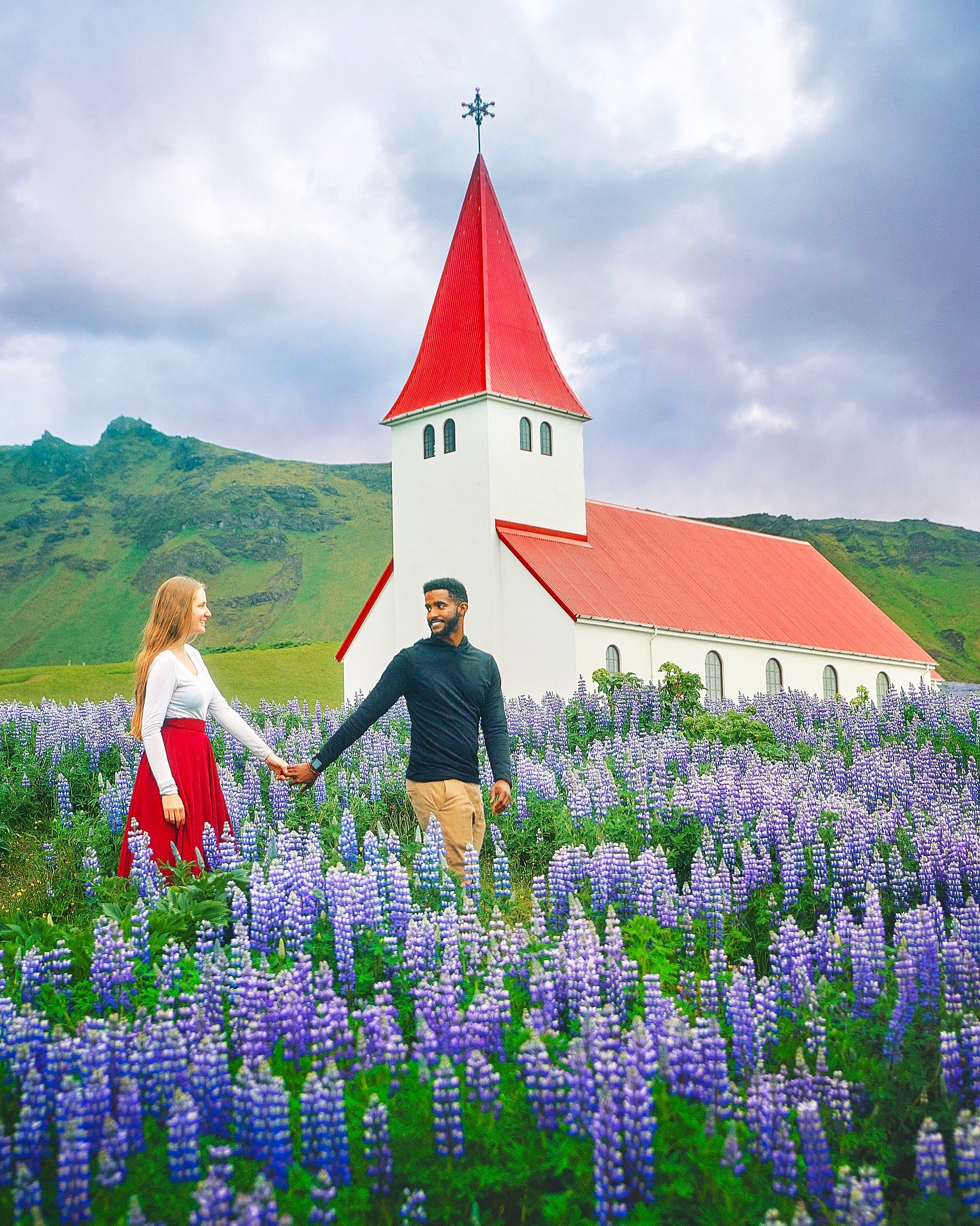 a woman and a man holding hands in a field of lupine flowers in front of a church, mountains in the background 