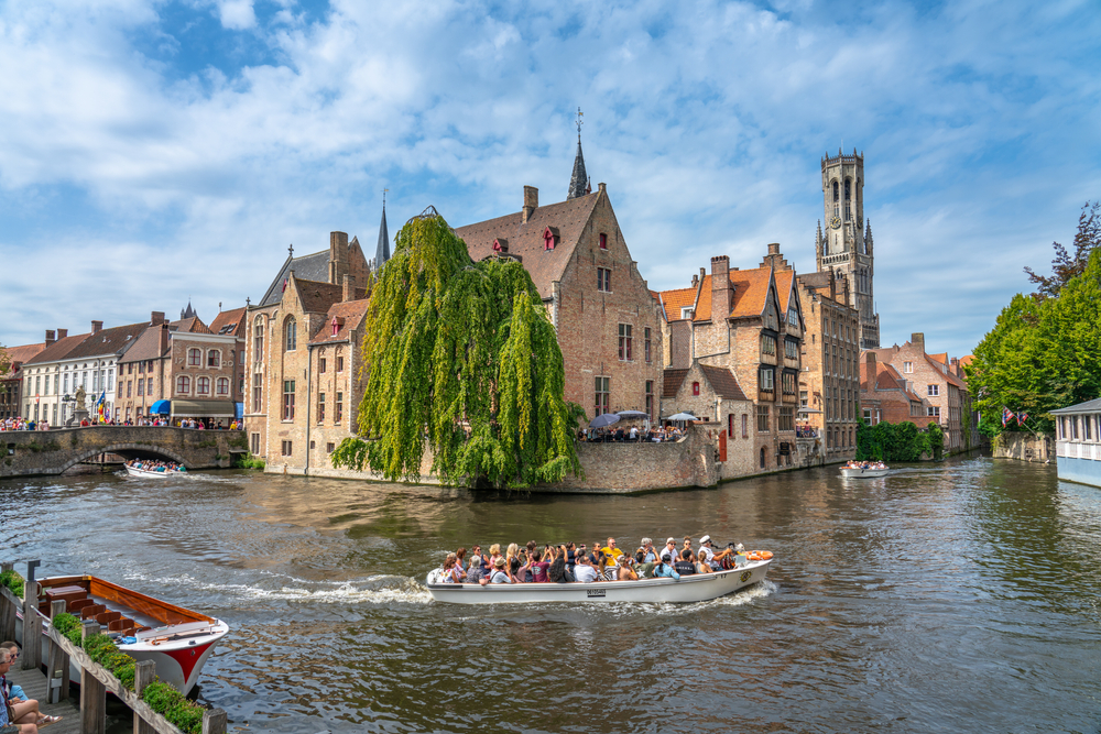 a boat full of people is going to the river in bruges belgium on a bright sunny day, buildings are along the river