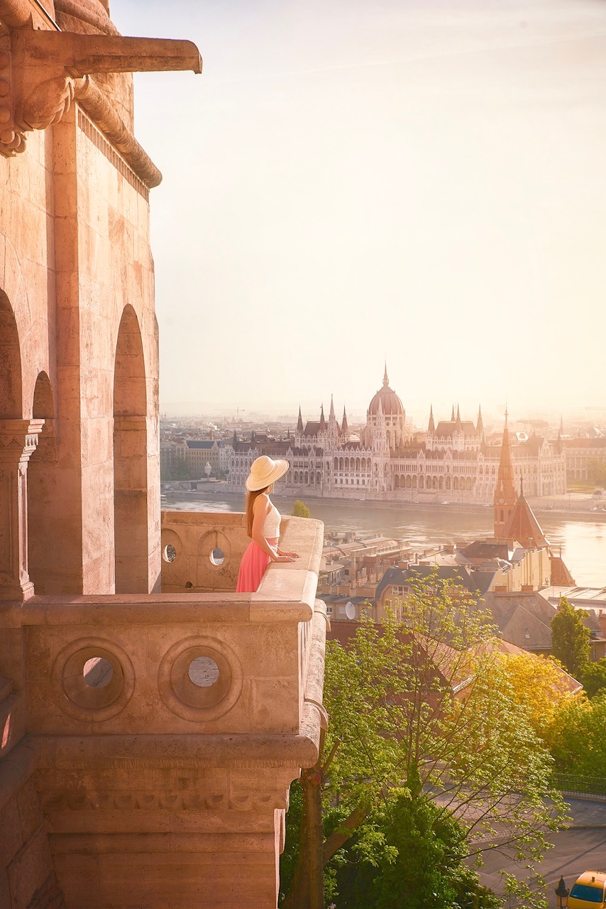 one of the best places to visit in europe in september, budapest. a woman is standing on a balcony in a large sun hat overlooking the river and the city 