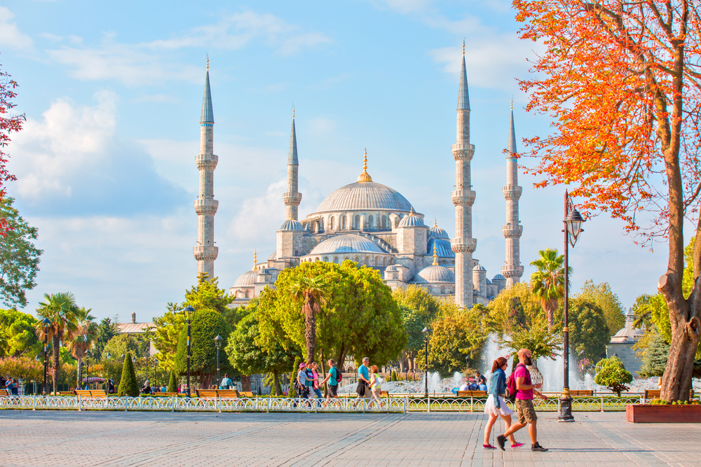 a grand mosque stands in the background with four tall towers, there is a fountain in front of it and people are walking around the fountain 