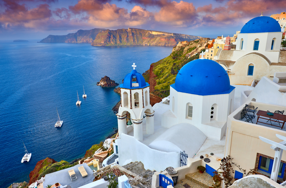 one of the best places to visit in europe in september, santorini greece during the day, ocean on the left, white and blue buildings on a cliff to the right 