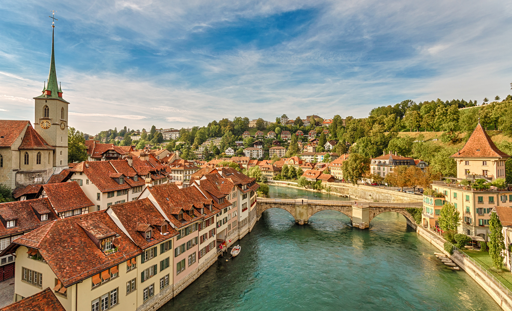 a swiss town, a tall clock tower is on the left, a river runs through the town and a bridge with three archways crosses it 