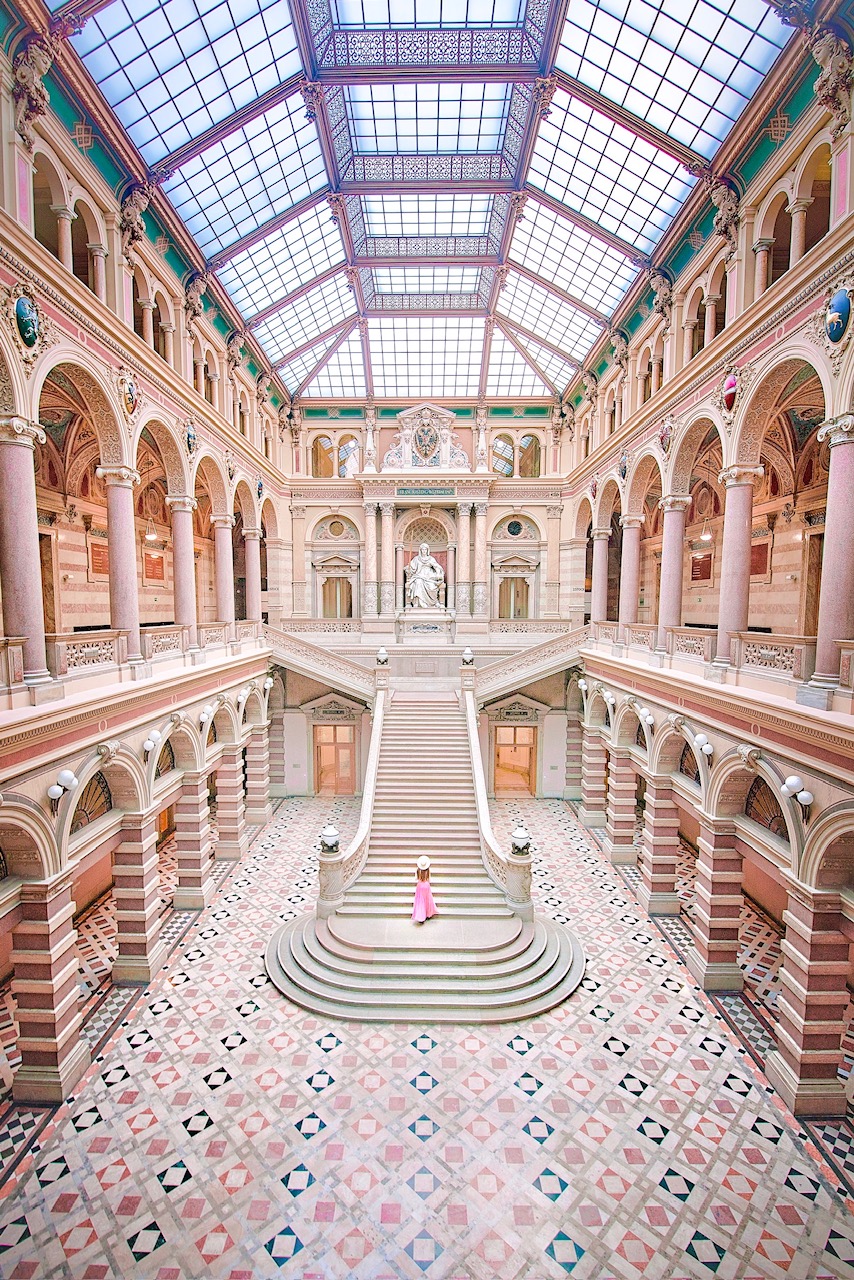 the ornate interior of a building. There are columns and archways on the sides of both stories and a long staircase in the middle of the image that leads between them, a woman is standing on the stairs. 
