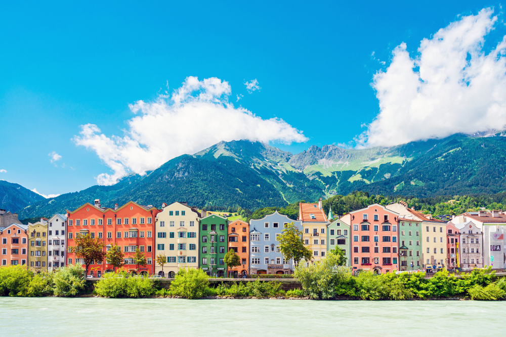 a row of tall colorful houses are lined up along the water in front of mountains on a partly cloudy day in Austria 