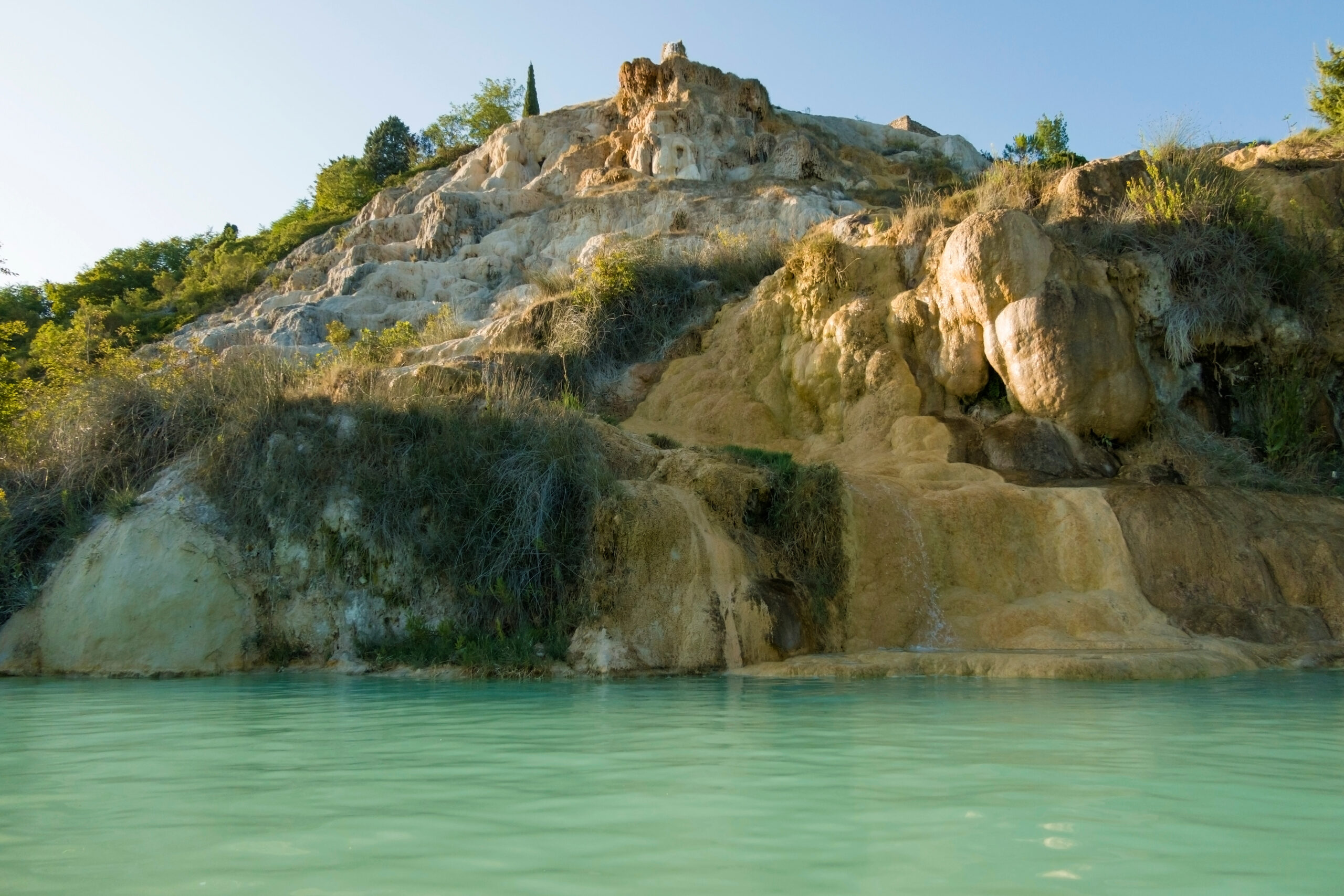 View of the natural hot springs at Parco Dei Mulini with a sandstone cliff face and jade green mineral rich waters 