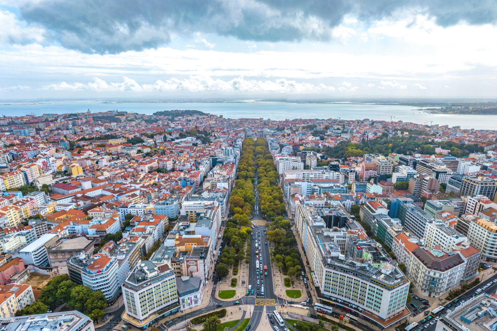 best places to visit in Europe in October, Lisbon  from above. a long road with trees on both sides can be seen in a city  with water in the background