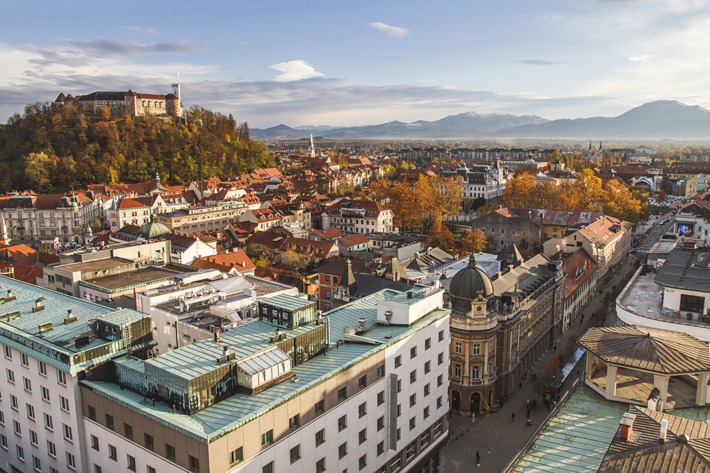 an aerial photo of a city in October, a building can be seen on a hill to the left and there are mountains in the background 