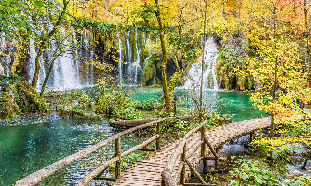 a wooden boardwalk curves over a lake with waterfalls falling into the water, Plitvice Lakes in October 