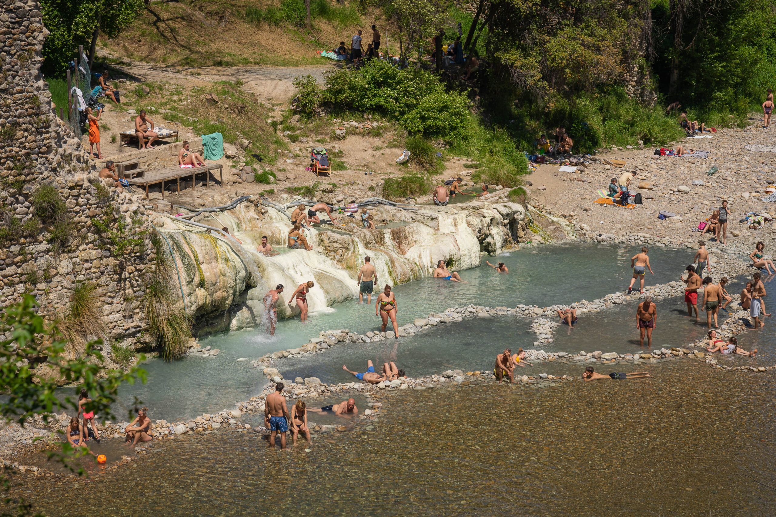 View of a crowd enjoying the Petriolo Hot Springs in Tuscany There are several pools a rocky beach and a bench visible. 