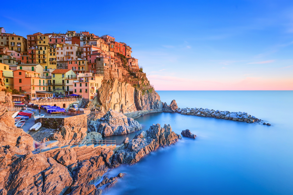 Manarola village on cliff rocks and sea at sunset. You can see a pink and blue sky and the houses and harbor. 