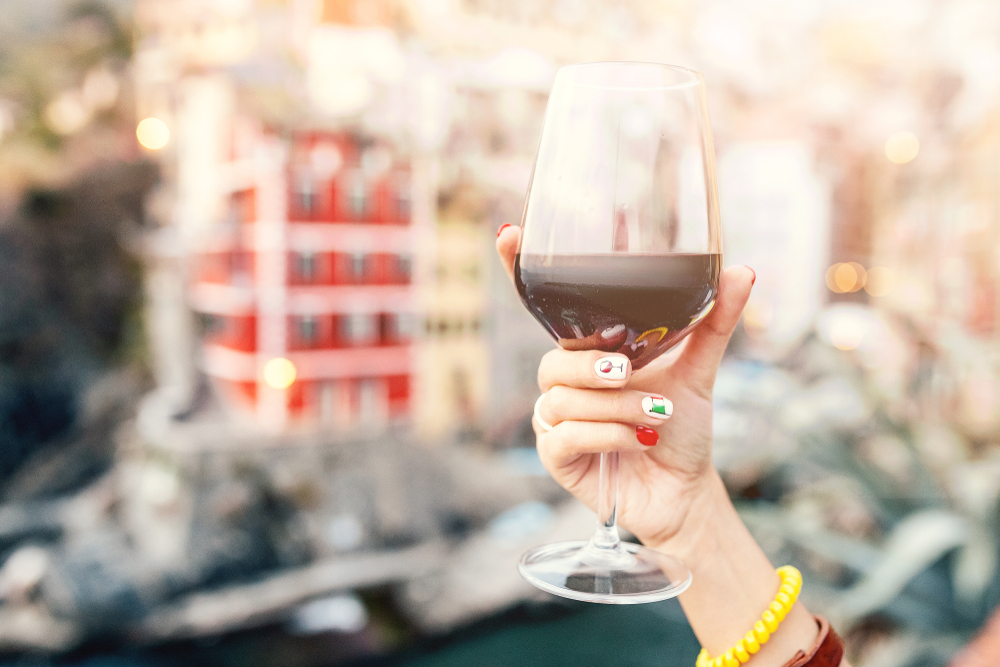 Female hand with italian flag nail art holding glass with red wine on a background of cinque terre riomaggiore town. Wine tasting is one of the things to do in Cinque Terre. 