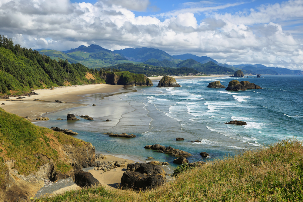 View looking down at the waves of Cannon Beach with sea stacks and rolling green hills in the distance during an Oregon road trip.
