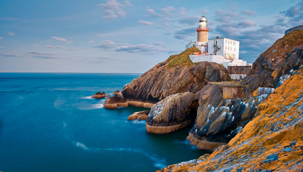 The Baily Lighthouse set on rugged brown cliffs along the ocean at sunset.