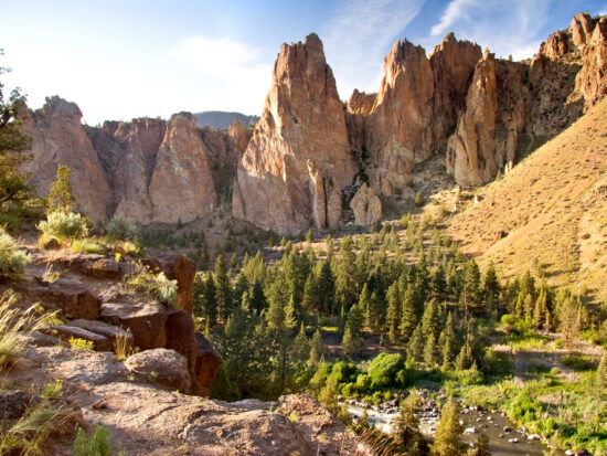 Sunny day over Smith Rock State Park with rugged rocks and a forest below.