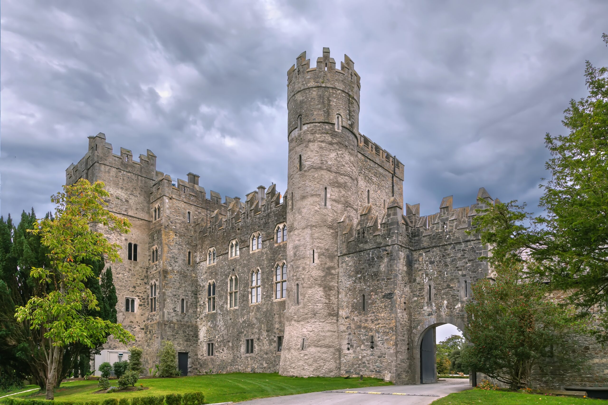 View of the drive of Kilkea Castle Hotel. It is surrounded by trees and greenery, made of beautiful grey stone and has gorgeous gothic windows. 