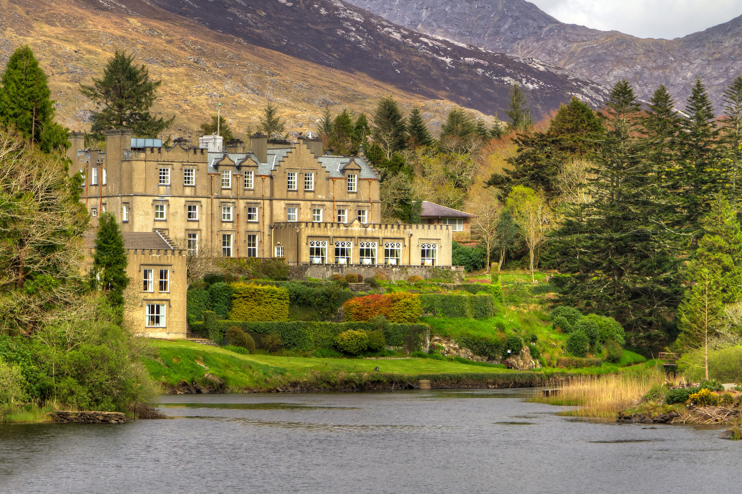 View of Ballynahinch Castle Hotel from the picturesque river including a view of the stunning mountains and evergreen trees surrounding it. 