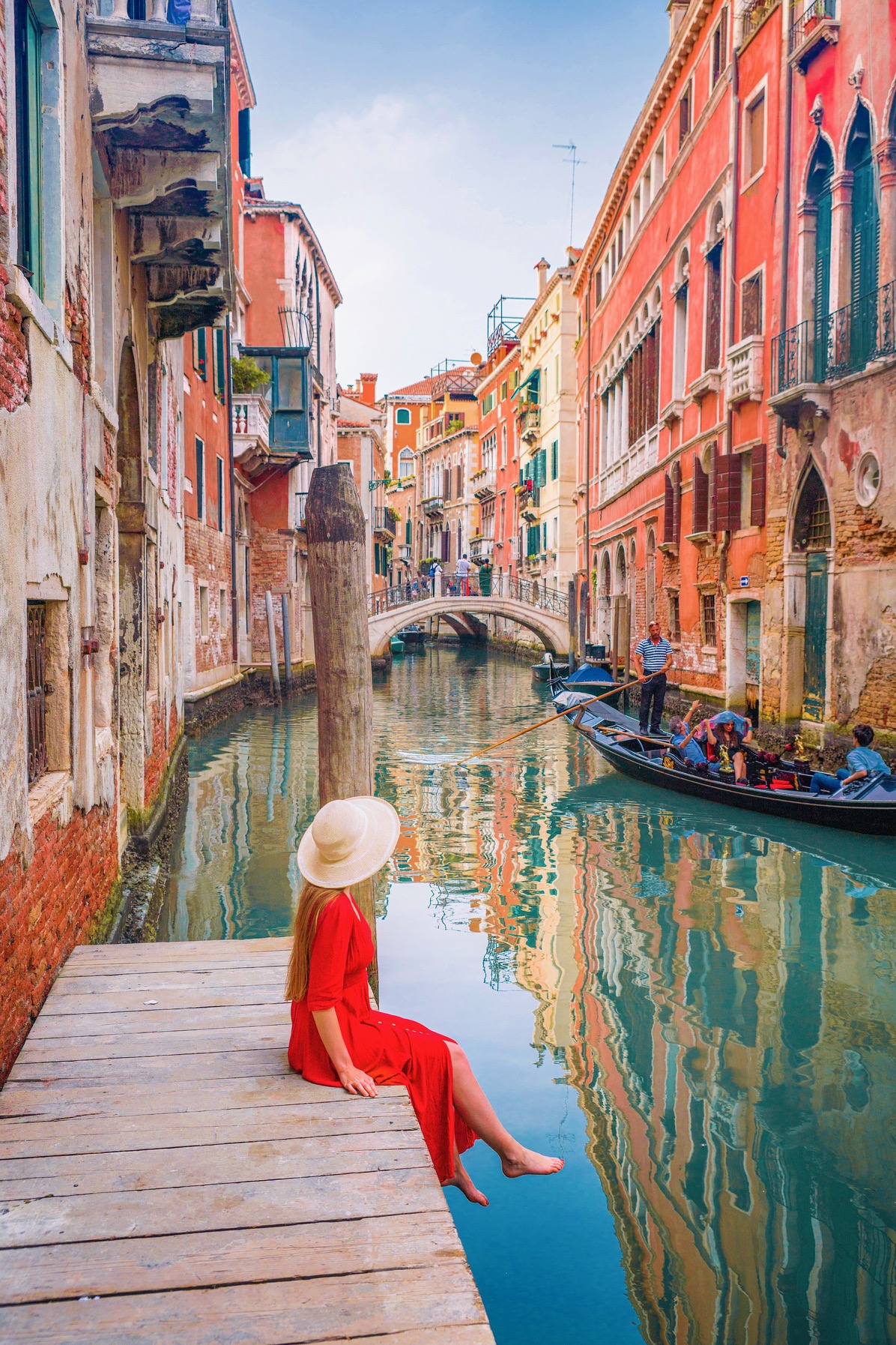 a girk in red dress and hat handing her feet over the dock on the canal as a gondola passes by