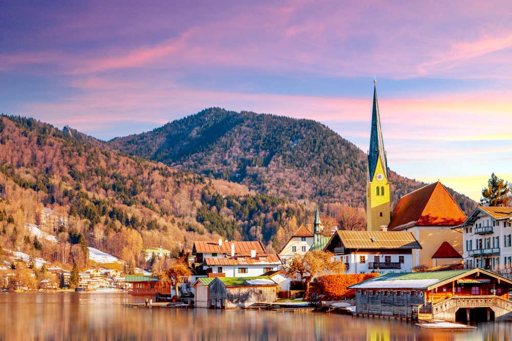 View over Rottach Egern. You can see the town by the lake with mountains in the background. You can see a church spire and old houses by the water. 