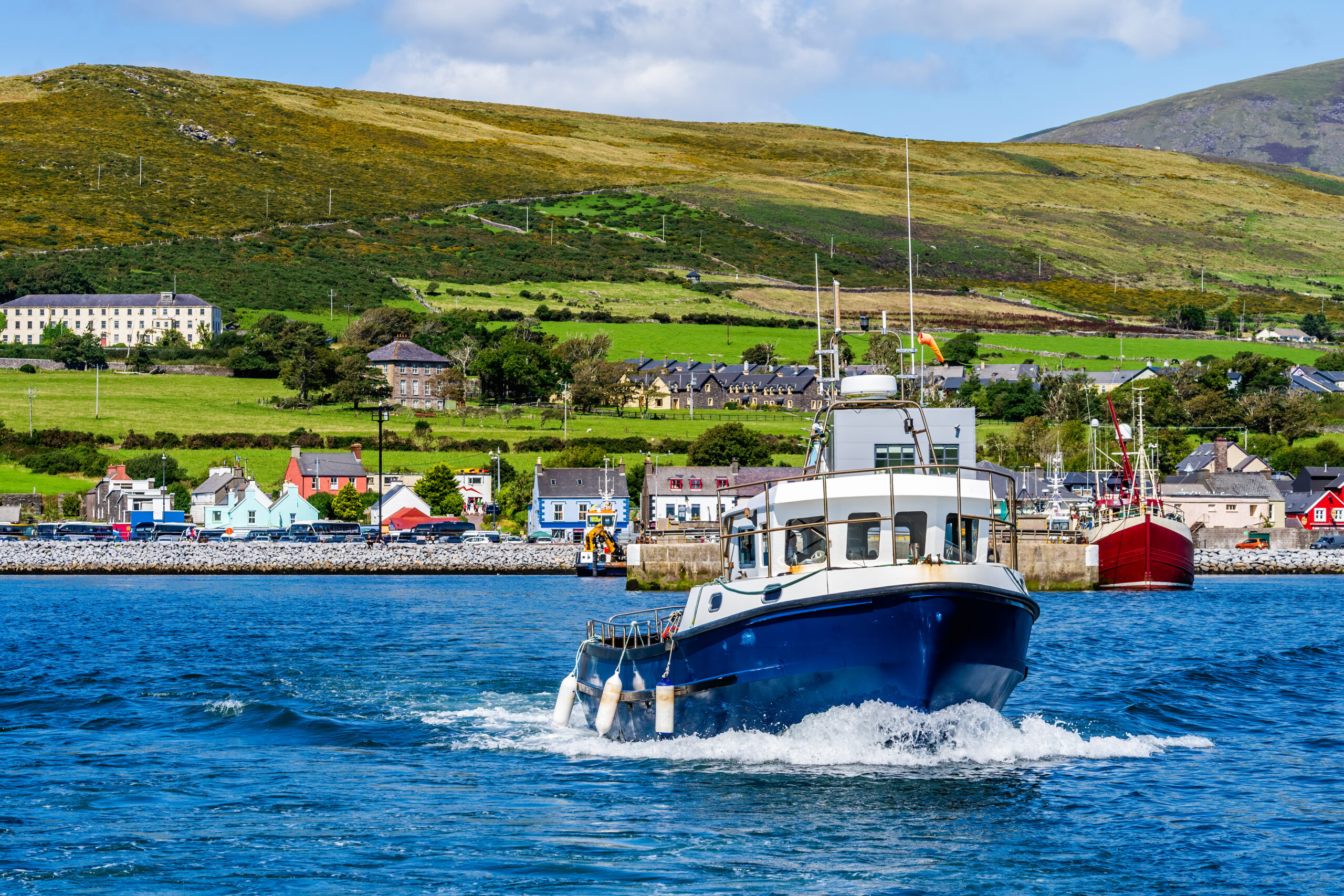 A small boat cutting through the deep blue waters of Dingle. A cute small town and rolling green hills are visible in the background. 