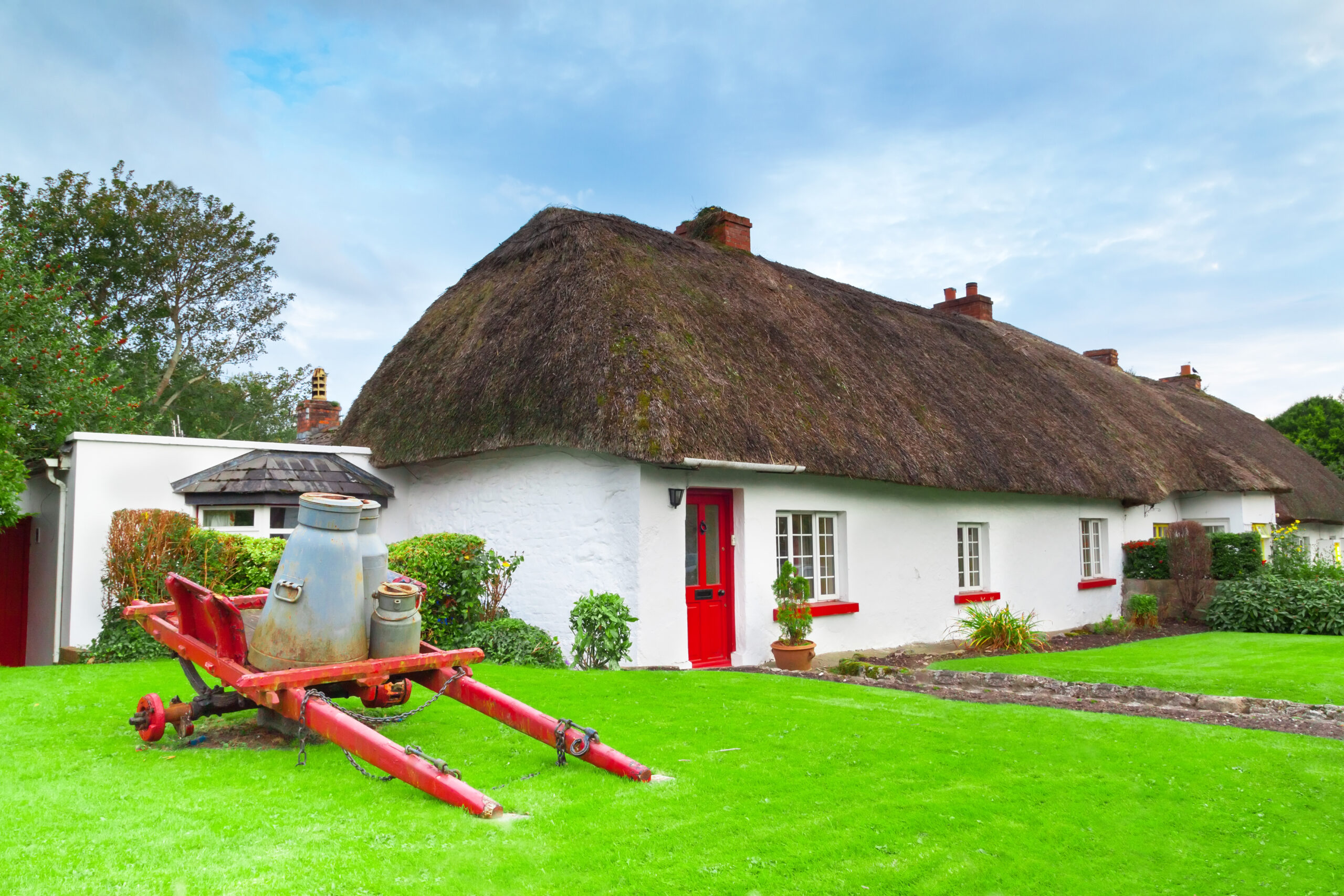 View of an old fashioned milk cart in front of a red and white vintage cottage with a thatched roof in Adare, Ireland.