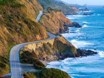 Aerial view of the Pacific Coast Highway in California along a rugged coastline with crashing waves during a West Coast road trip.