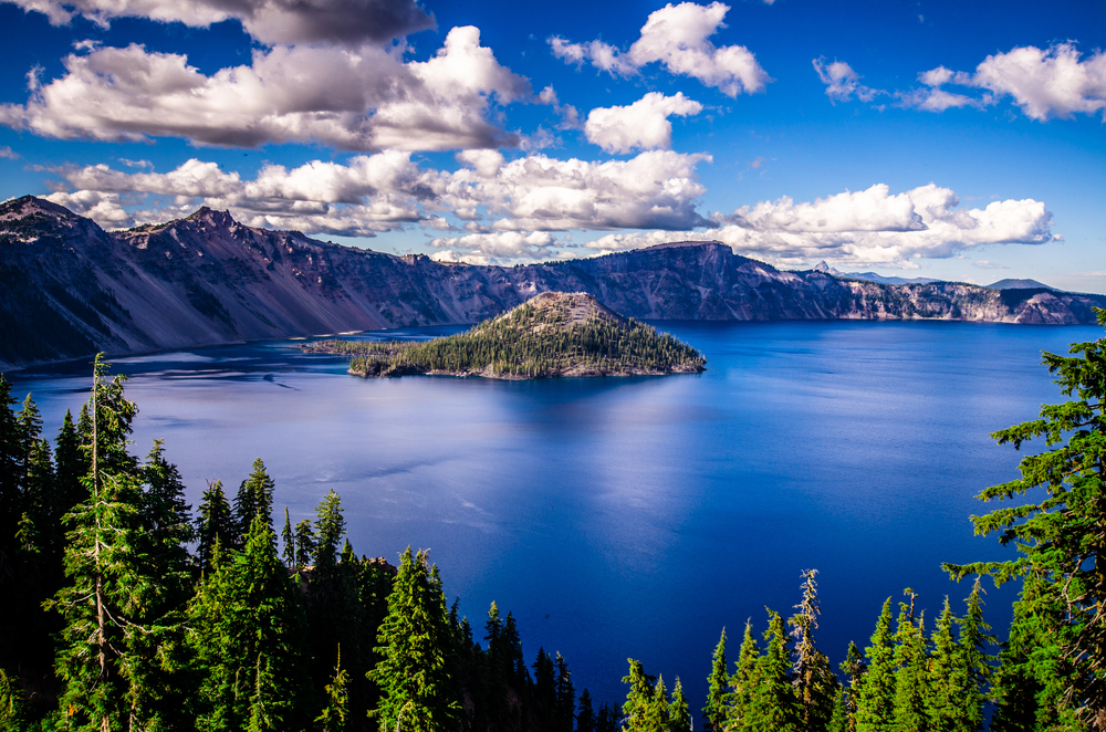 View looking down at Crater Lake with deep blue water and an island. There are mountains on the other side and a smattering of clouds overhead.