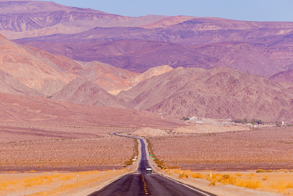 Road running through the desert of Death Valley National Park with pink and purple colored mountains on a  West Coast road trip.