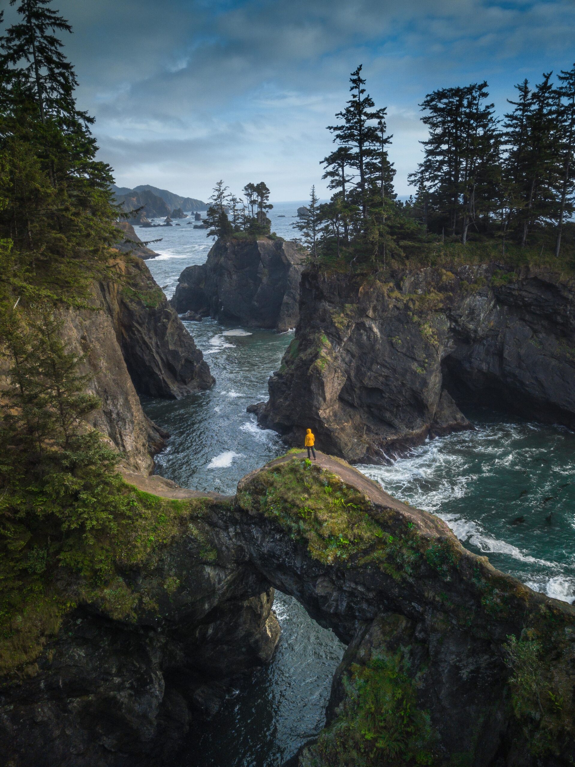 A figure in a yellow rain jacket standing on a natural arch over looking the ocean on the Samuel H. Boardman State Scenic Corridor in Oregon on an overcast day.
