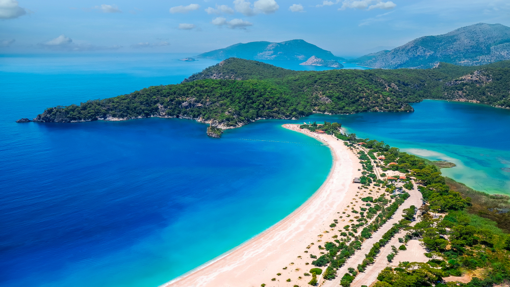 View of Ölüdeniz Beach from above. The water is varying shades of turquoise and blue, the sand is white, and the surrounding vegetation is very green. 