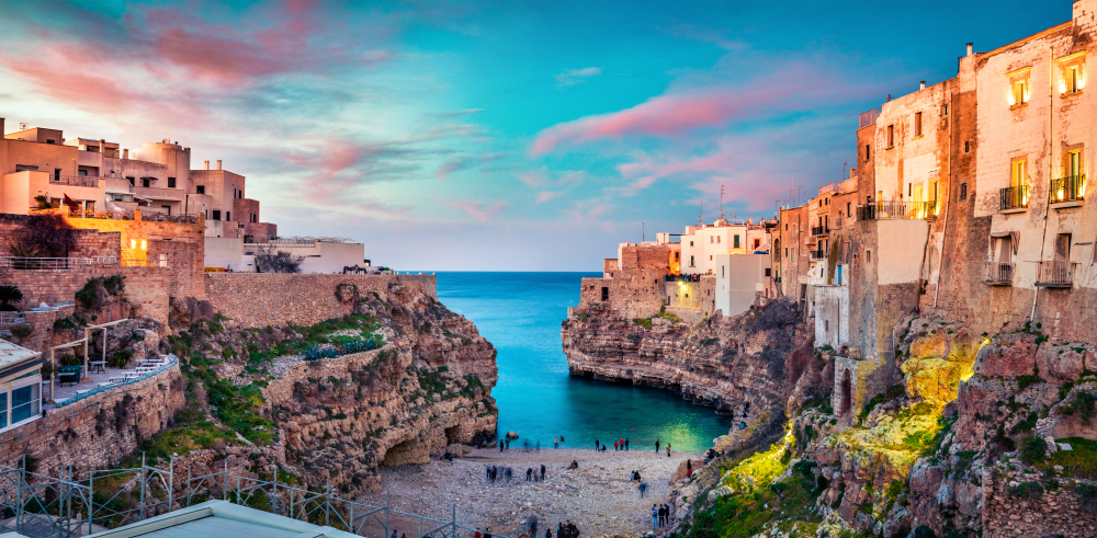 View of Polignano a Mare Beach during sunset with crystal blue water, pink sunset clouds, and orange light hitting the buildings surrounding one of the best beaches in Europe.