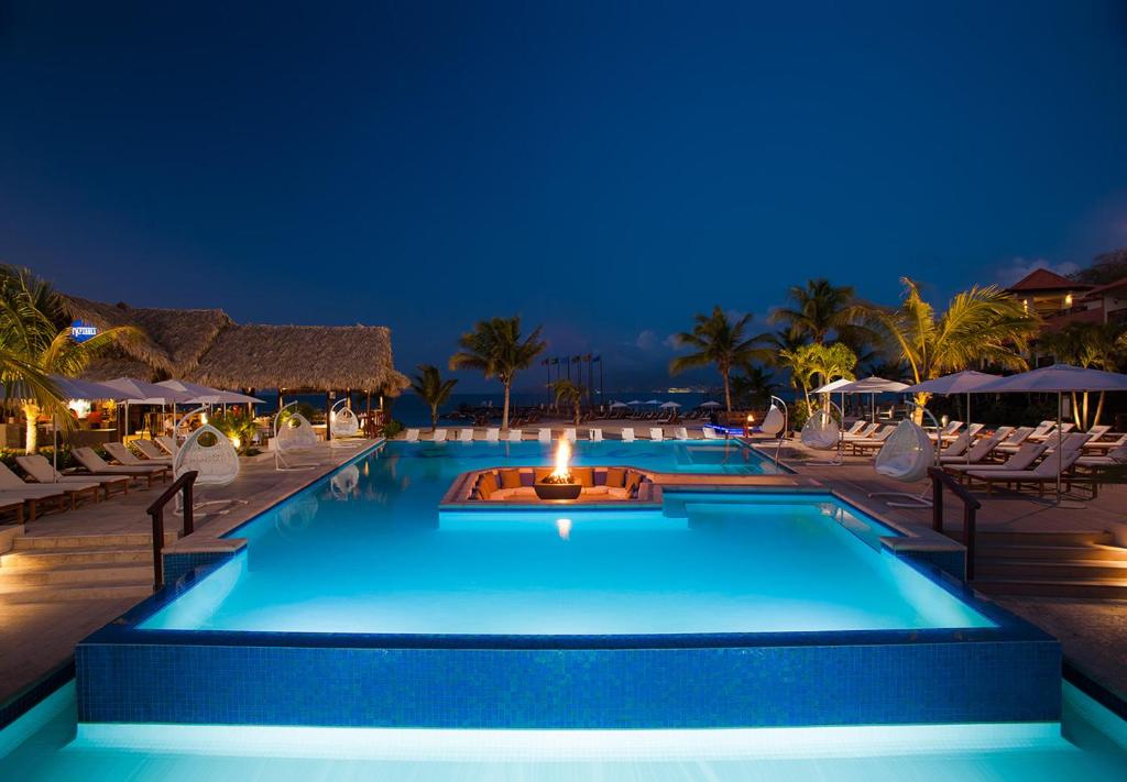 Hotel pool surrounded by lunge chairs. There is a seating area and fire pit in the middle and you can see the ocean in the distance. 