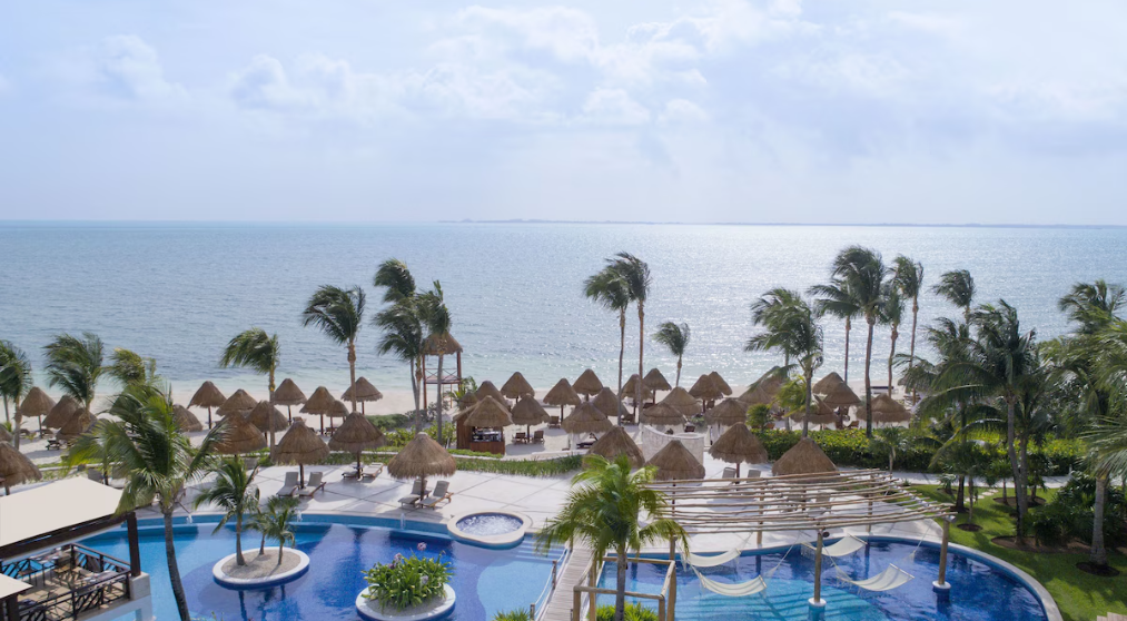 Hotel pool surrounded by straw umbrellas with the sea in the background. 