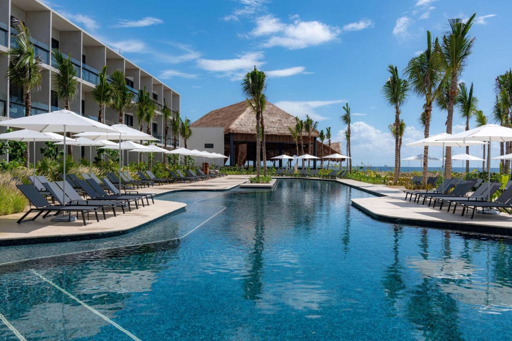 View of a pool with a thatch roof building in the background. There are chairs and umbrellas around the pool. 