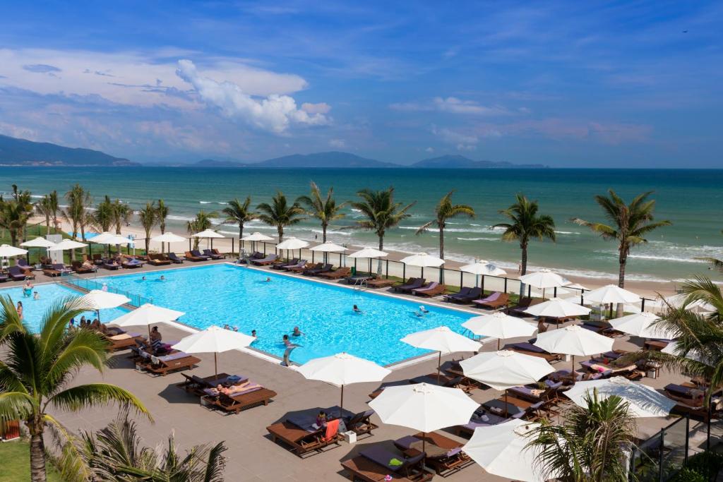 Swimming pool surrounded by umbrellas and palm trees. It is on the beachfront and you can see the sea and distance mountains. 