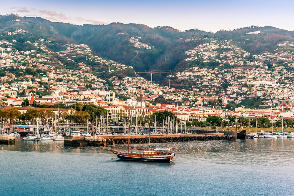 A view of the marina and city of Funchal, Maderia on a sunny morning. You can see a boat in the foreground and the town in the background. 