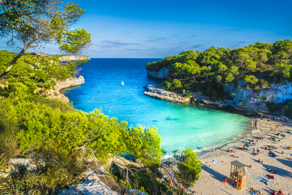 Aerial view of Cala Llombards, one of the best beaches in Europe. The crystal clear, blue green water leads to a white sandy beach with people scattered about. The cove is surrounded by small cliffs topped with green trees. 