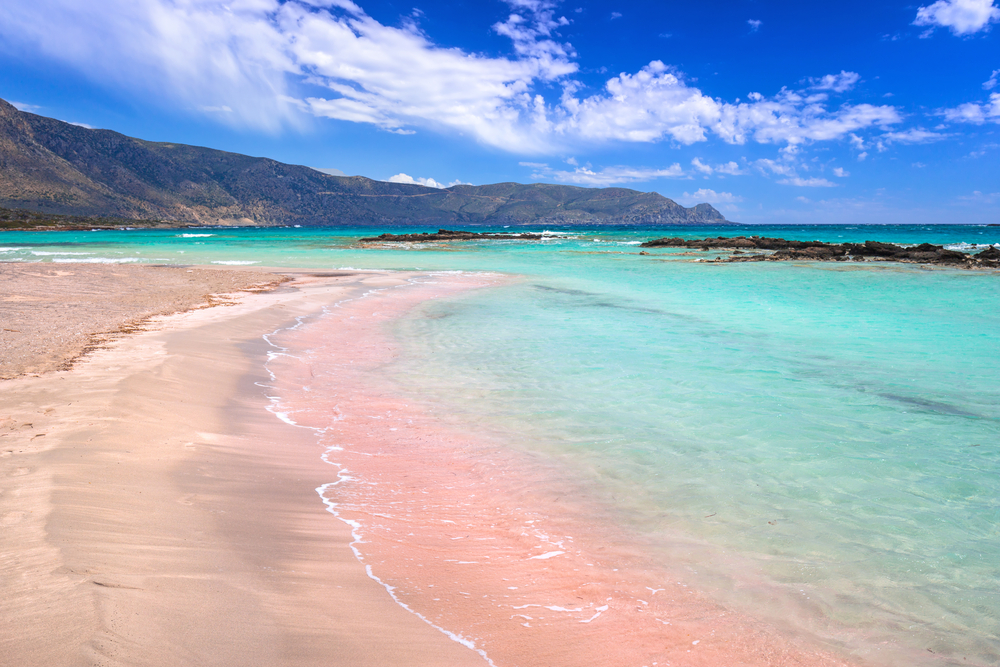 Crystal clear blue waters lap against the shore filled with pink sand at Elafonisi Beach in Crete.